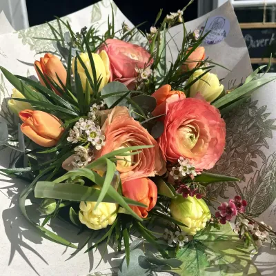 Bouquet de renoncules corail, tulipes orange et jaunes, wax flower et feuillages fins dans un papier imprimé