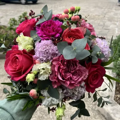 Bouquet de roses fuchsia et rouges, œillets violets, lisianthus blancs, millepertuis et eucalyptus tenu à la main