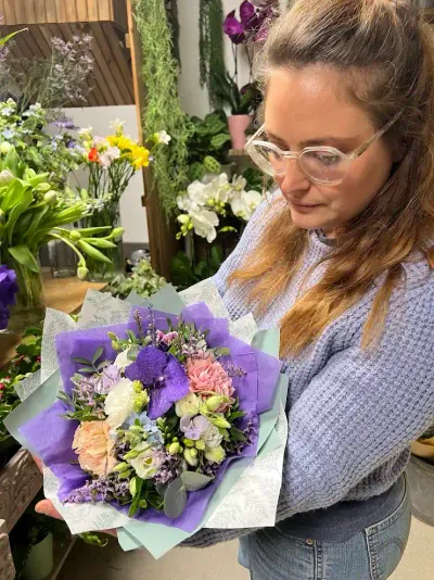 Léa Mahé regarde un bouquet d'orchidées vanda, roses pêche et lisianthus dans un papier violet, en boutique