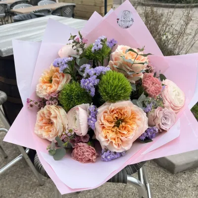 Bouquet de roses pêche, œillets boules verts, statice mauve et œillets anciens dans un papier rose, en terrasse