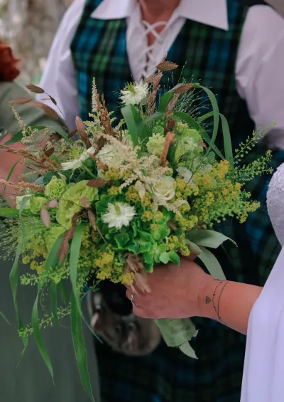 Bouquet de mariée champêtre vert et blanc avec astilbe, nigelles et graminées, tenu devant un gilet écossais