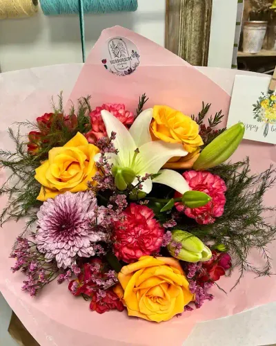 Bouquet de lys blanc, roses jaunes, œillets rouges et chrysanthème mauve dans un papier rose étiqueté Herborium