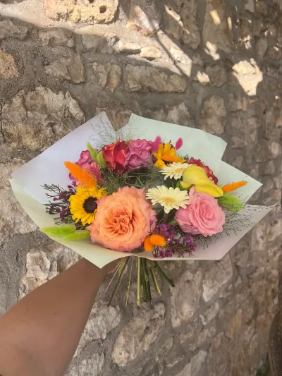Bouquet coloré de rose pêche, tournesol, gerbera crème, calla jaune et queues de lièvre orange, devant un mur en pierre