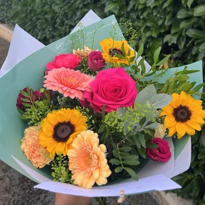 Bouquet coloré de tournesols, gerberas pêche et roses fuchsia, emballé dans un papier vert d'eau