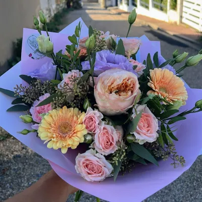 Bouquet pastel de roses de jardin pêche, gerberas abricot et lisianthus mauves, dans un papier lilas