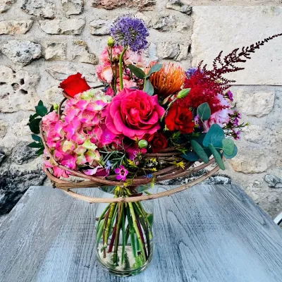 Bouquet structuré en vase : hortensia rose, roses rouges, alliums et protéa, devant un mur de pierre