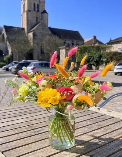 Bouquet en vase de fleurs jaunes et orange et de queues de lièvre teintées, sur une table devant une église