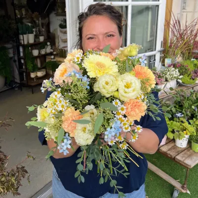 Bouquet pastel jaune et pêche de gerberas, roses et petites fleurs bleues, tenu par Léa dans la boutique Herborium
