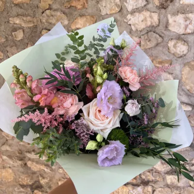 Bouquet pastel de lisianthus mauves, roses crème, œillets roses et mufliers, devant un mur de pierre