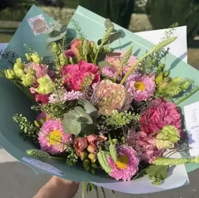 Bouquet de roses et œillets roses, asters, lisianthus et graminées dans un papier vert d'eau, tenu à la main