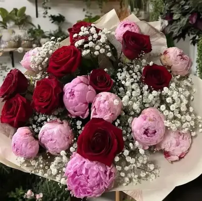 Grand bouquet de pivoines roses, roses rouges et gypsophile dans un papier crème, devant les plantes de la boutique