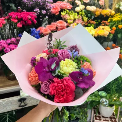 Bouquet coloré d'orchidées violettes, œillets et roses mauves dans un papier rose, devant les fleurs de la boutique Herborium