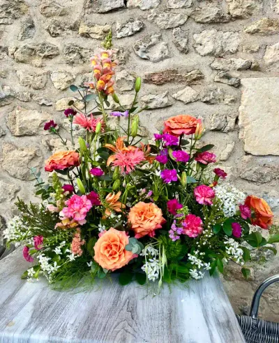 Devant de cercueil haut aux couleurs vives, roses orange, œillets fuchsia, gerberas et mufliers, devant un mur de pierre