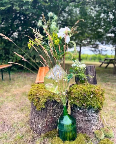 Décor de mariage champêtre : souches couvertes de mousse, dame-jeanne verte, vase de nigelles et herbes folles
