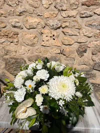Composition de deuil blanche : chrysanthème boule, anthurium, œillets, marguerites et roses blanches, devant un mur de pierre