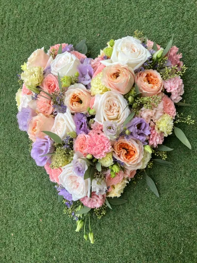 Cœur de deuil en roses de jardin pêche et blanches, lisianthus mauves et œillets roses, posé à plat sur un gazon