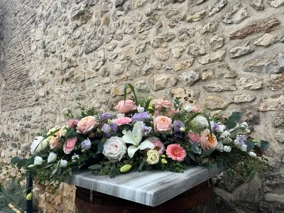 Dessus de cercueil pastel : roses de jardin pêche, lys blanc, gerbera rose et lisianthus mauves, devant un mur de pierre