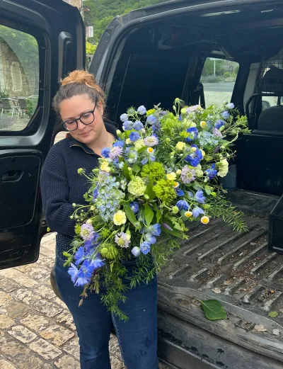 Léa Mahé porte une grande raquette de deuil en delphiniums bleus, fleurs blanches et feuillages, près de sa camionnette