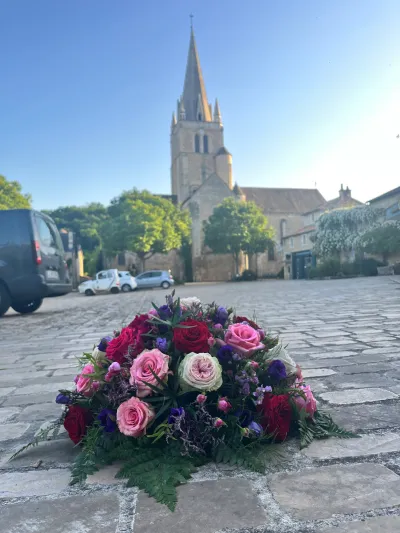 Coussin de deuil rond en roses rouges et roses, lisianthus violets et fougère, posé sur les pavés devant une église