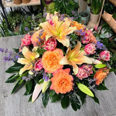 Coussin de deuil rond aux lys orangés, roses orange, tulipes roses et lavande, sur une table de la boutique