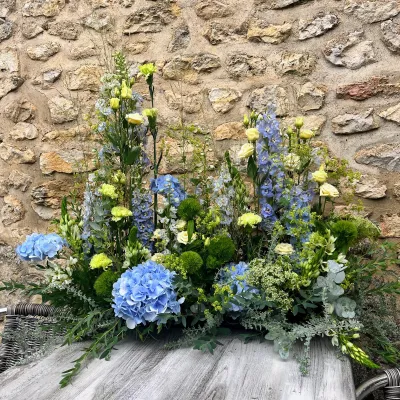 Devant de cercueil champêtre bleu : delphiniums, hortensia bleu et fleurs vert tendre, devant un mur de pierre