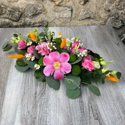 Raquette de deuil aux pivoines roses, œillets, gerberas et ammi, posée sur une table en bois gris
