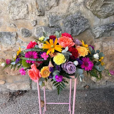 Raquette de deuil colorée sur un pied rose, rubans de feuilles écrits « À notre Maman » et « À notre Mémé »