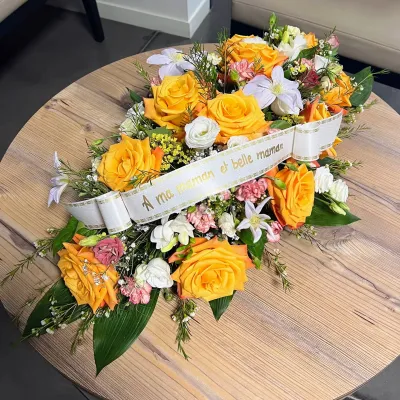 Raquette de deuil aux roses orange et fleurs blanches, ruban « À ma maman et belle maman », sur une table