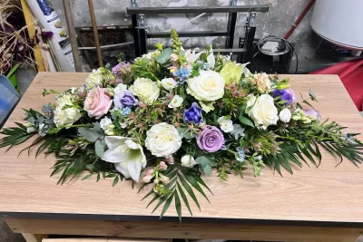 Raquette de deuil blanche aux roses blanches, lys, lisianthus violets et palmes, posée sur une table