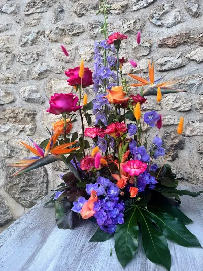 Composition de deuil haute et colorée : oiseaux de paradis, delphiniums, roses fuchsia, devant la pierre