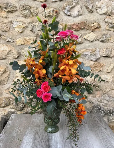 Composition florale haute dans un vase vert : orchidées orange, roses fuchsia et eucalyptus, devant la pierre
