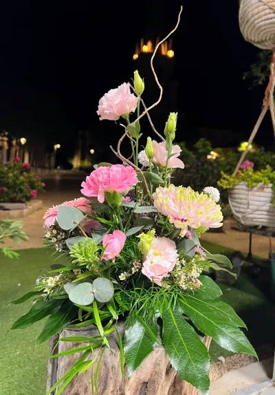 Composition florale d'œillets roses, lisianthus et grandes feuilles sur un rondin, dehors à la nuit tombée