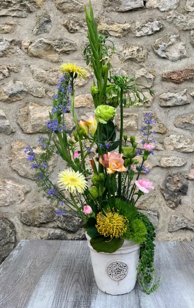 Composition haute dans un pot blanc : gerberas jaunes, lisianthus, fleurs bleues et verdure, devant la pierre