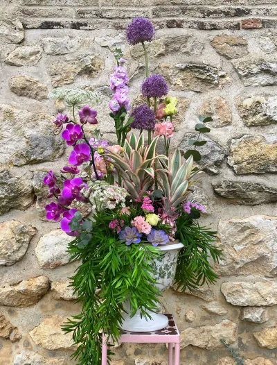 Grande composition en vasque sur un pied rose : orchidées violettes, alliums, roses et feuillage retombant
