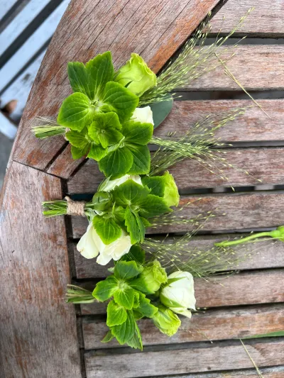 Trois boutonnières de mariage : clochettes d'Irlande vertes, bouton blanc et graminées, sur une table en bois