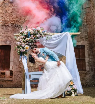 Mariés s'embrassant sous une arche fleurie pastel, fumigènes roses, bleus et verts, dans des ruines en pierre