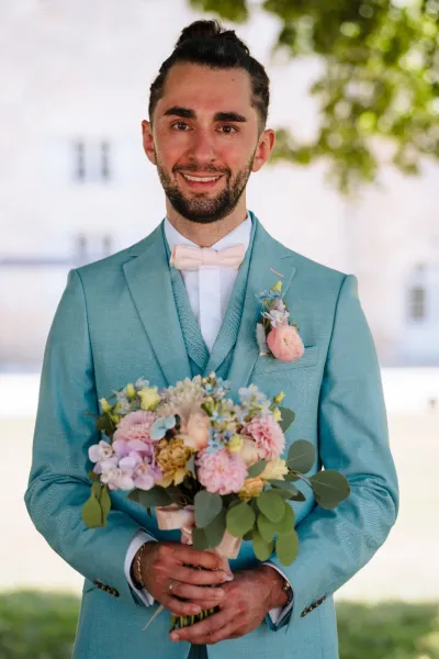 Marié en costume turquoise tenant le bouquet de mariée pastel, avec une boutonnière de rose pêche assortie