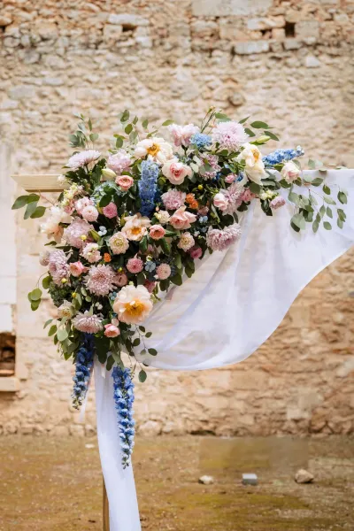 Angle d'arche de mariage en roses pêche, dahlias et delphiniums bleus avec voile blanc, contre un mur de pierre