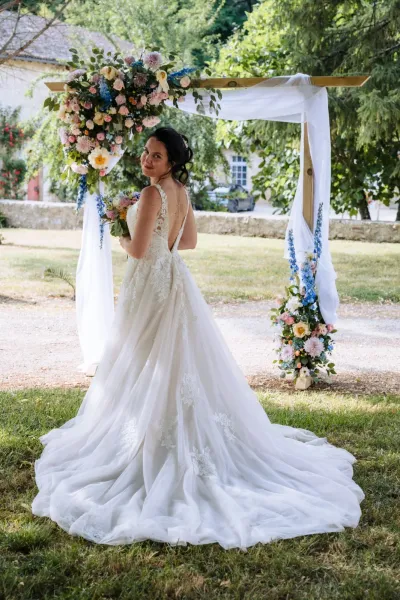 Mariée de dos, robe à longue traîne, devant une arche de mariage fleurie de roses et de delphiniums bleus