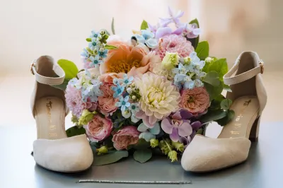 Bouquet de mariée pastel en dahlias, roses et petites fleurs bleues, posé entre les escarpins de la mariée