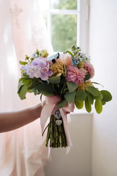 Bouquet de mariée pastel tenu devant une fenêtre lumineuse, roses et dahlias parmi l'eucalyptus, ruban blanc