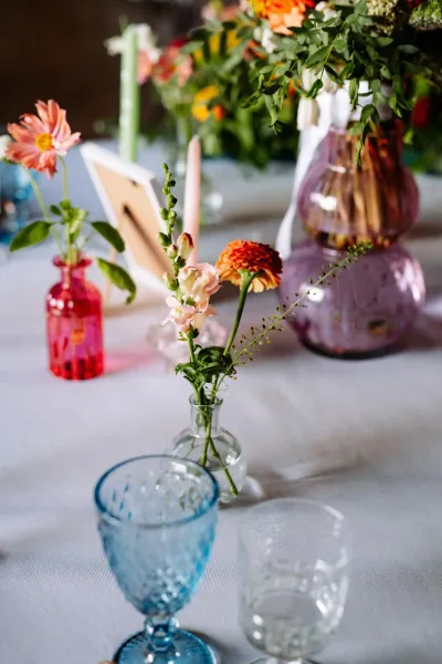 Table de mariage : soliflores de fleurs roses et de dahlias orange dans des fioles colorées, verres à pied bleus