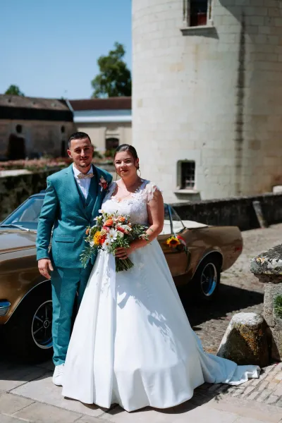 Mariés devant une voiture ancienne et une tour de château, la mariée tient un bouquet de fleurs orange et roses