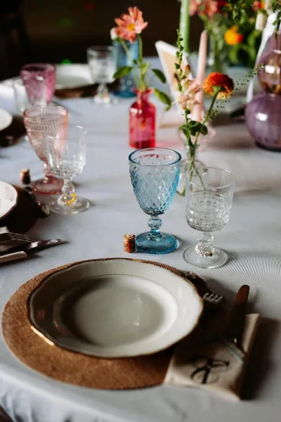 Couvert de mariage : assiette dorée, verres à pied bleus et fioles de fleurs roses et orange sur la nappe blanche