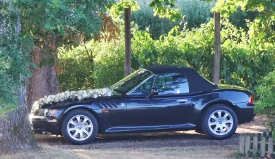 Cabriolet noir des mariés décoré d'une guirlande de roses blanches et de gypsophile sur le capot, garé sous les arbres