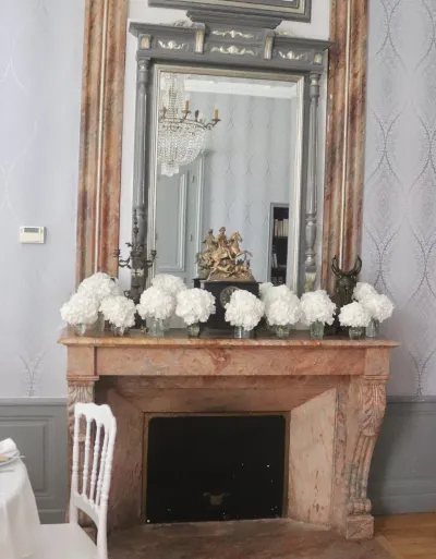 Petits vases d'hortensias blancs alignés sur une cheminée en marbre sous un grand miroir, décoration florale d'un mariage