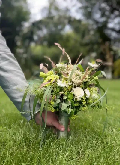 Main tenant un bouquet de mariée champêtre de roses blanches, lisianthus et graminées, au ras de l'herbe