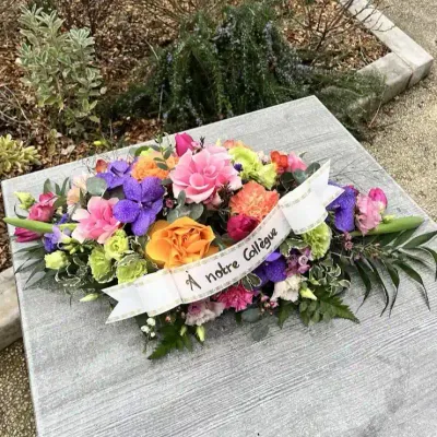 Raquette de deuil colorée de roses, œillets orange et orchidées vanda, avec un ruban « À notre collègue »