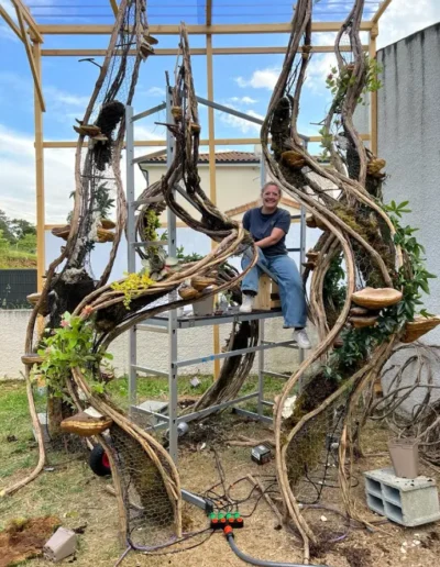 Lea Mahe posant a cote de la structure metallique et du grillage de sa scenographie vegetale Peoch en pleine creation