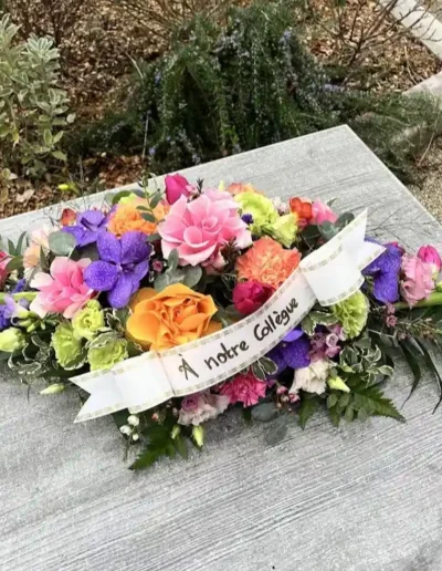 Raquette de fleurs allongee couleurs vives avec ruban blanc marque A notre Collegue posee sur un support pierre